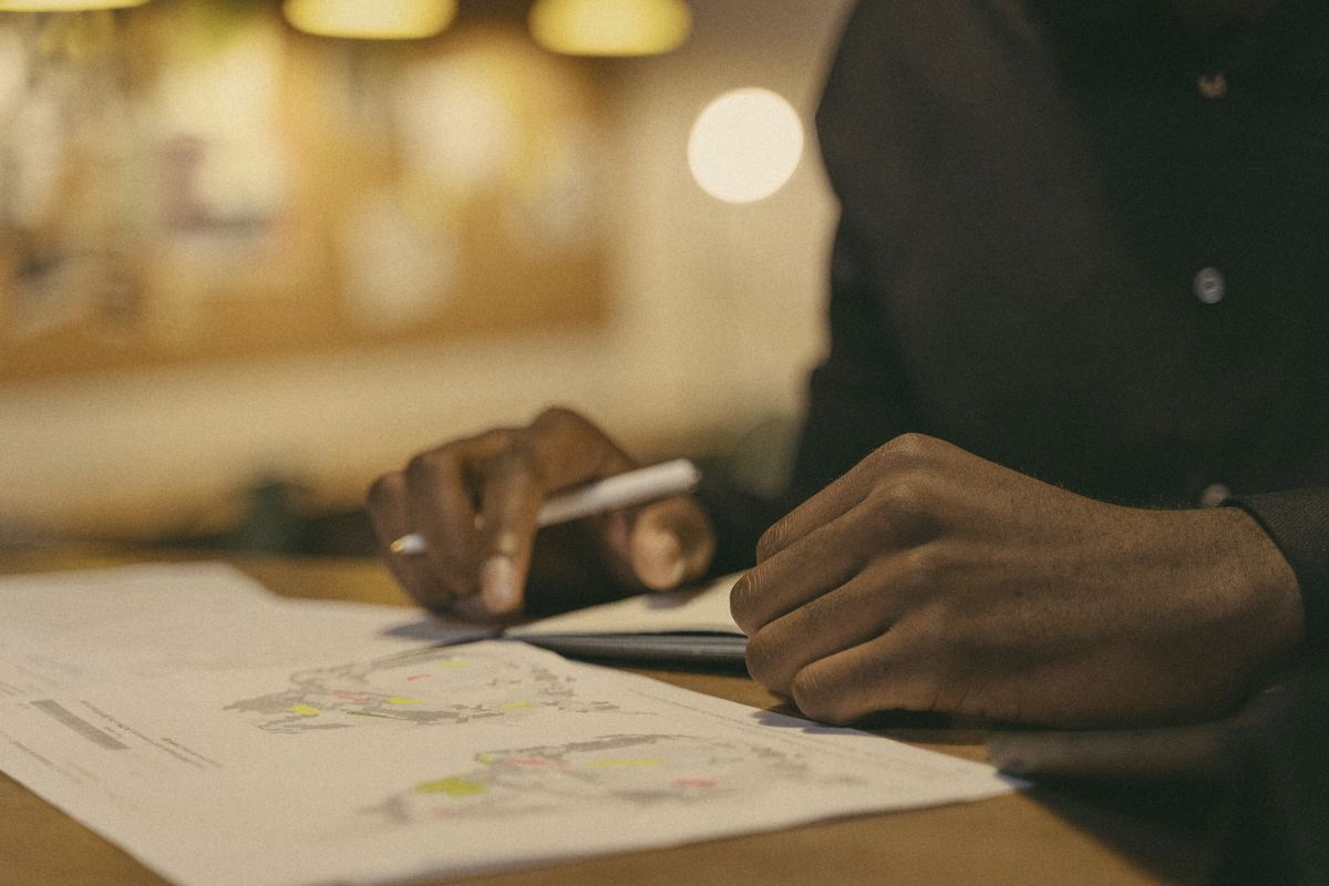 Hands holding a pen over a printed map on a wooden table in warm lamplight