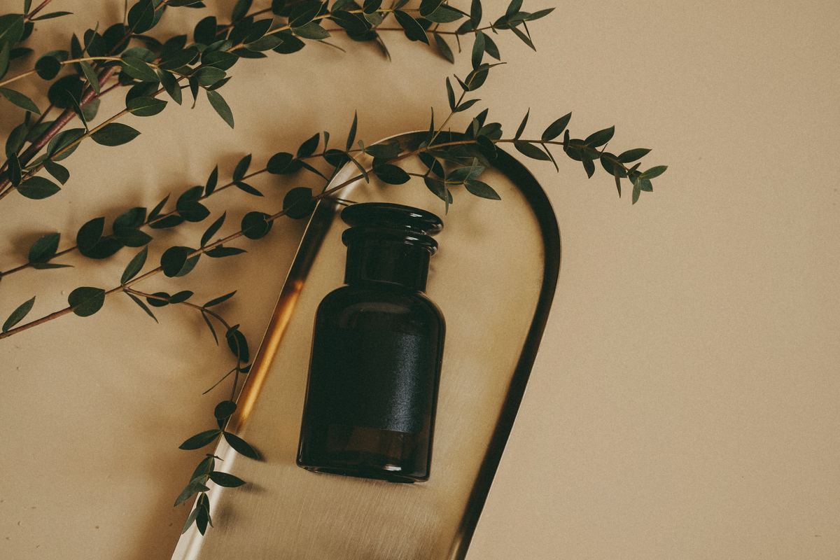 Unlabelled amber glass jar on a brass tray with sprigs of eucalyptus on a warm sand background