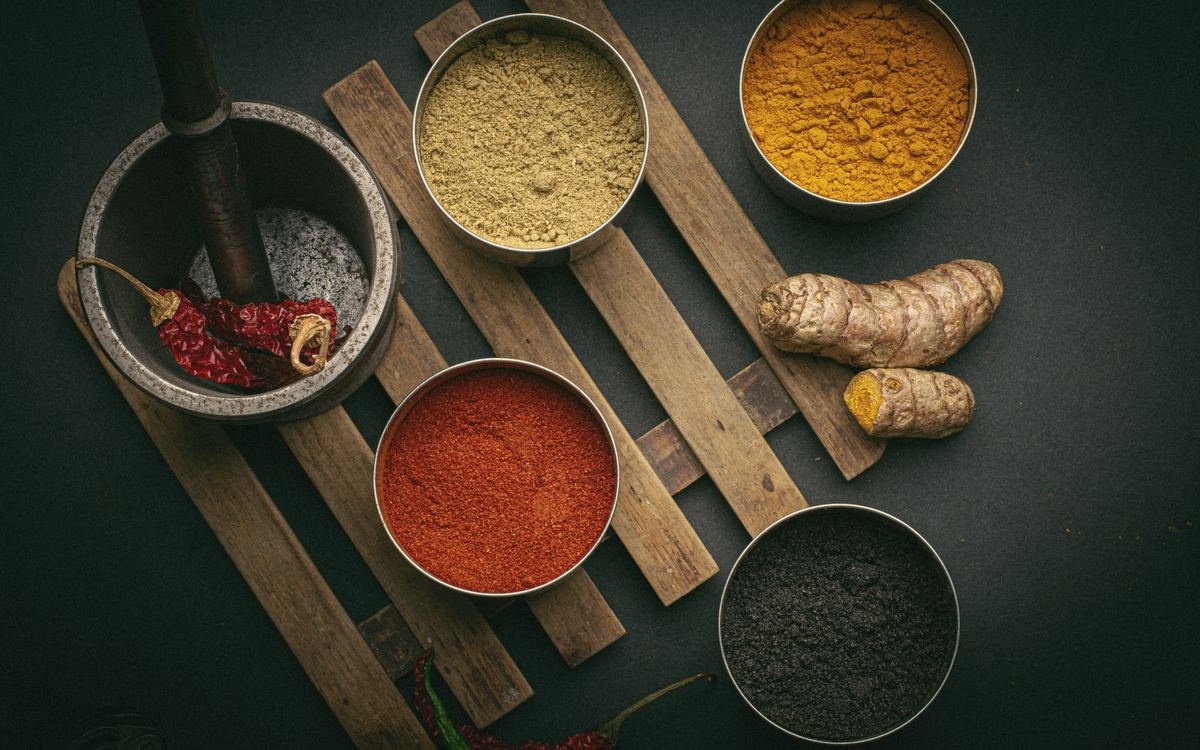 Overhead view of tins of ground spices, fresh turmeric and dried chillies on a dark wooden board